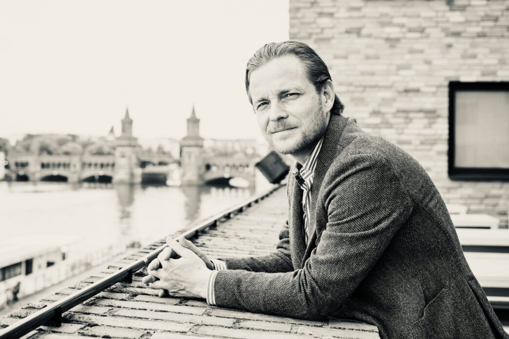 Portrait of a man in a blazer leaning on a railing on a rooftop with a distant cityscape in the background (black and white).