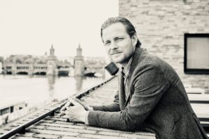Portrait of a man in a blazer leaning on a railing on a rooftop with a distant cityscape in the background (black and white).