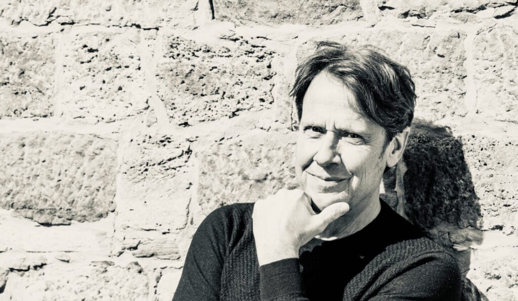 Man leaning against a rough stone wall, hand resting on chin in a thoughtful pose.