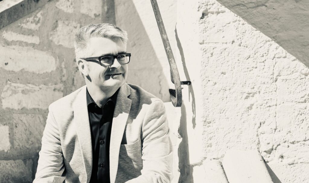Man in a light blazer and black shirt sits on stone stairs, looking to the side, wearing glasses.