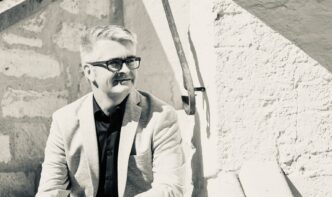 Man in a light blazer and black shirt sits on stone stairs, looking to the side, wearing glasses.