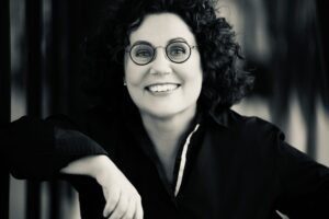 Smiling woman with curly hair and round glasses, looking at the camera in black and white.