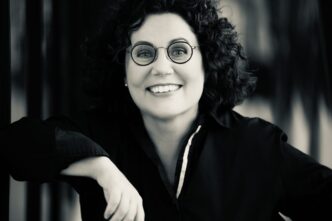 Smiling woman with curly hair and round glasses, looking at the camera in black and white.