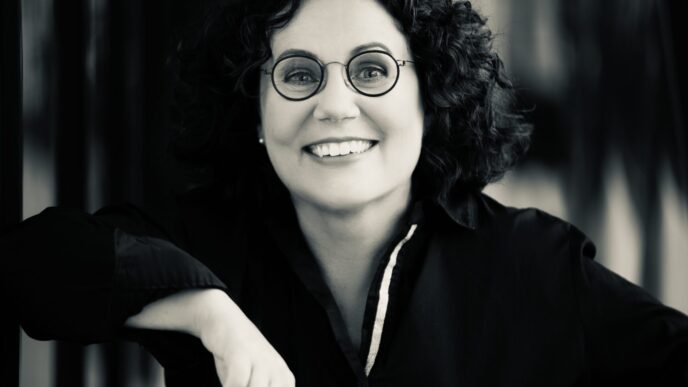 Smiling woman with curly hair and round glasses, looking at the camera in black and white.