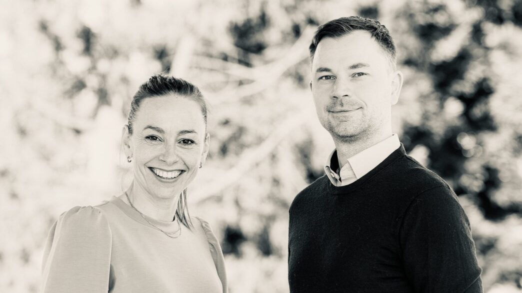 Smiling woman on the left and man on the right posing outdoors with a blurred natural background.