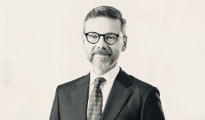 Professional headshot of a man in a dark suit and patterned tie, wearing round black glasses.