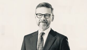 Professional headshot of a man in a dark suit and patterned tie, wearing round black glasses.