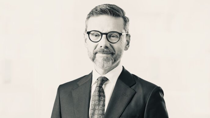 Professional headshot of a man in a dark suit and patterned tie, wearing round black glasses.