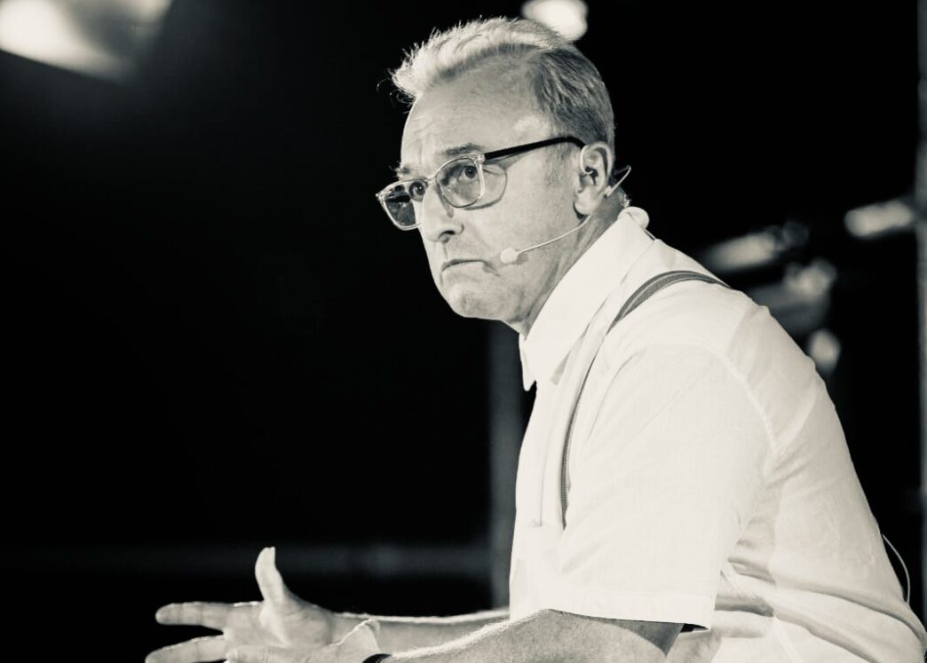 Male presenter on stage wearing a headset microphone, glasses, white shirt, and suspenders, gesturing with open hands as he speaks.