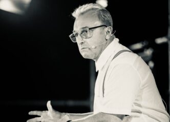 Male presenter on stage wearing a headset microphone, glasses, white shirt, and suspenders, gesturing with open hands as he speaks.