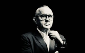 Portrait of an older man in a suit with glasses, posed thoughtfully with a hand near his chin against a black background.