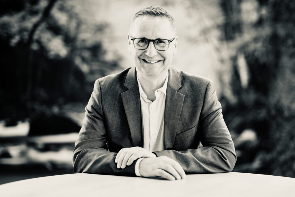 Smiling man in a blazer and glasses seated at a table outdoors, black-and-white photo.