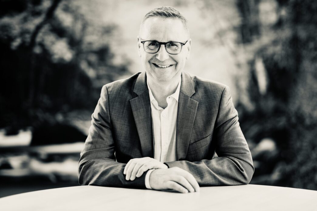Smiling man in a blazer and glasses seated at a table outdoors, black-and-white photo.