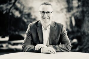 Smiling man in a blazer and glasses seated at a table outdoors, black-and-white photo.