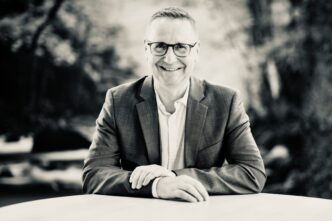 Smiling man in a blazer and glasses seated at a table outdoors, black-and-white photo.