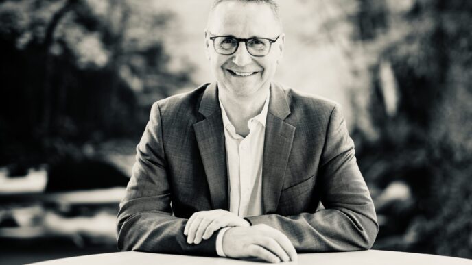 Smiling man in a blazer and glasses seated at a table outdoors, black-and-white photo.