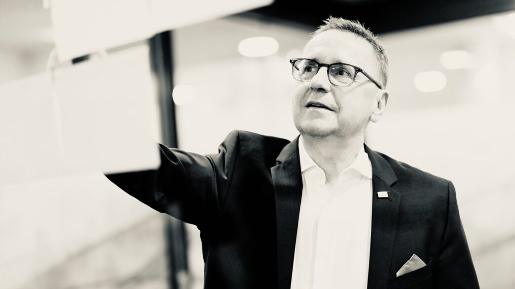 Businessman in a suit and glasses reaches forward, mid-presentation in a modern office setting.