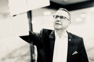 Businessman in a suit and glasses reaches forward, mid-presentation in a modern office setting.
