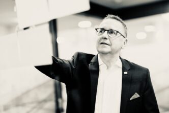 Businessman in a suit and glasses reaches forward, mid-presentation in a modern office setting.