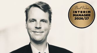 Black-and-white portrait of a smiling man in a suit with a gold circular badge reading 'Best Value Interim Manager 2026/27' on the right.