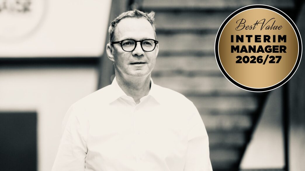 Man wearing glasses in a white shirt stands next to a gold badge reading 'Best Value Interim Manager 2026/27' on a black-and-white background,