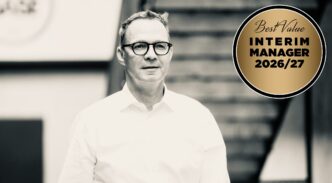 Man wearing glasses in a white shirt stands next to a gold badge reading 'Best Value Interim Manager 2026/27' on a black-and-white background,