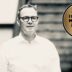 Man wearing glasses in a white shirt stands next to a gold badge reading 'Best Value Interim Manager 2026/27' on a black-and-white background,