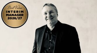 Smiling man in a black suit beside a gold circular badge that says 'Best Value Interim Manager 2026/27'.