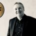 Smiling man in a black suit beside a gold circular badge that says 'Best Value Interim Manager 2026/27'.
