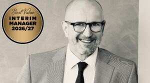 Portrait of a smiling man in a gray suit and glasses, with a gold circular badge reading 'Best Value Interim Manager 2026/27' on the left side.