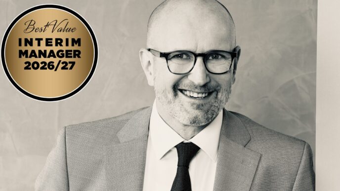 Portrait of a smiling man in a gray suit and glasses, with a gold circular badge reading 'Best Value Interim Manager 2026/27' on the left side.