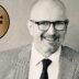 Portrait of a smiling man in a gray suit and glasses, with a gold circular badge reading 'Best Value Interim Manager 2026/27' on the left side.