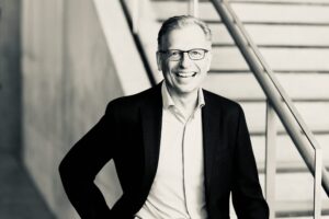 Smiling man in a suit and glasses, standing near a stair railing (black-and-white photo).